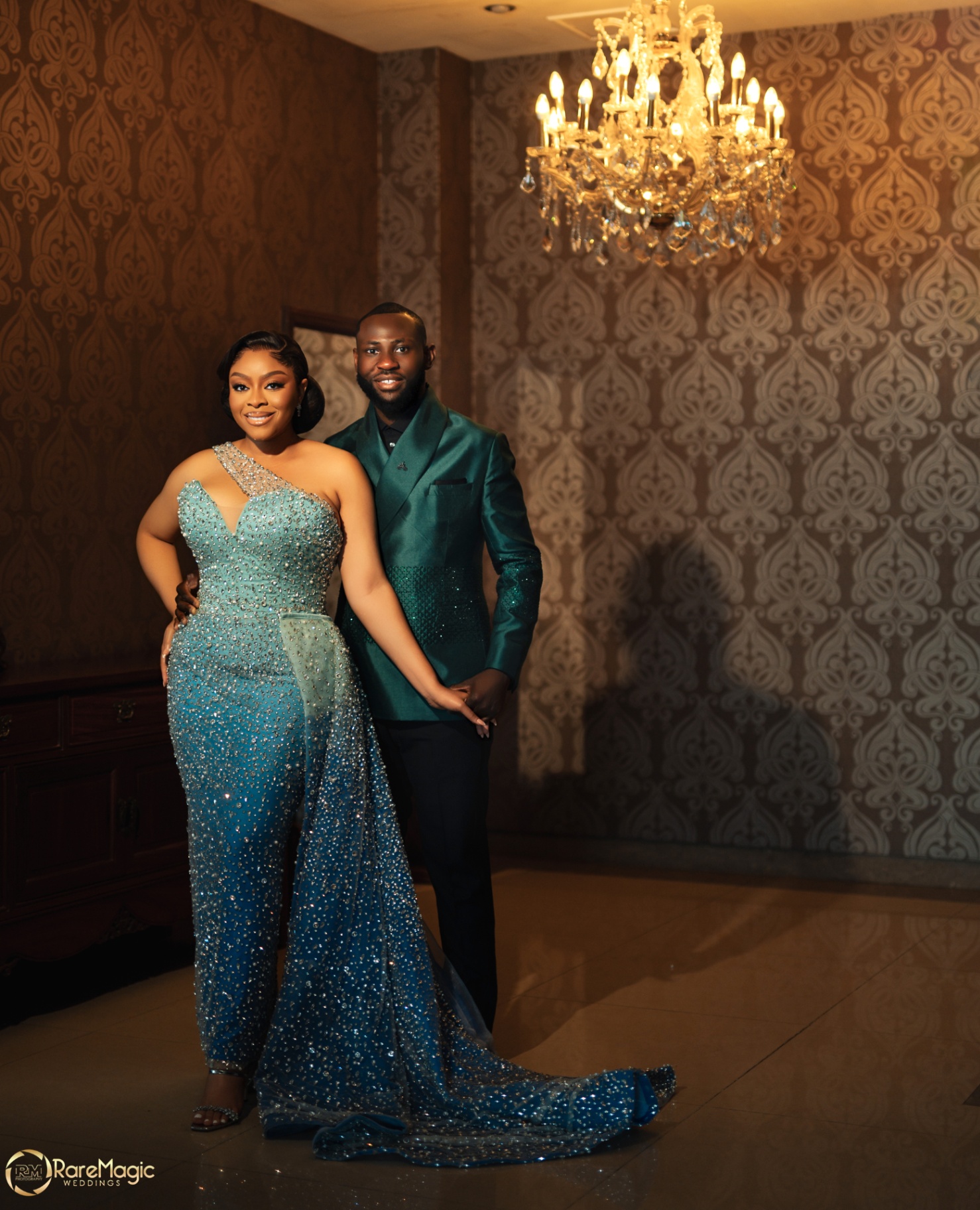 Couple portrait in teal beaded gown and emerald jacket beneath a chandelier