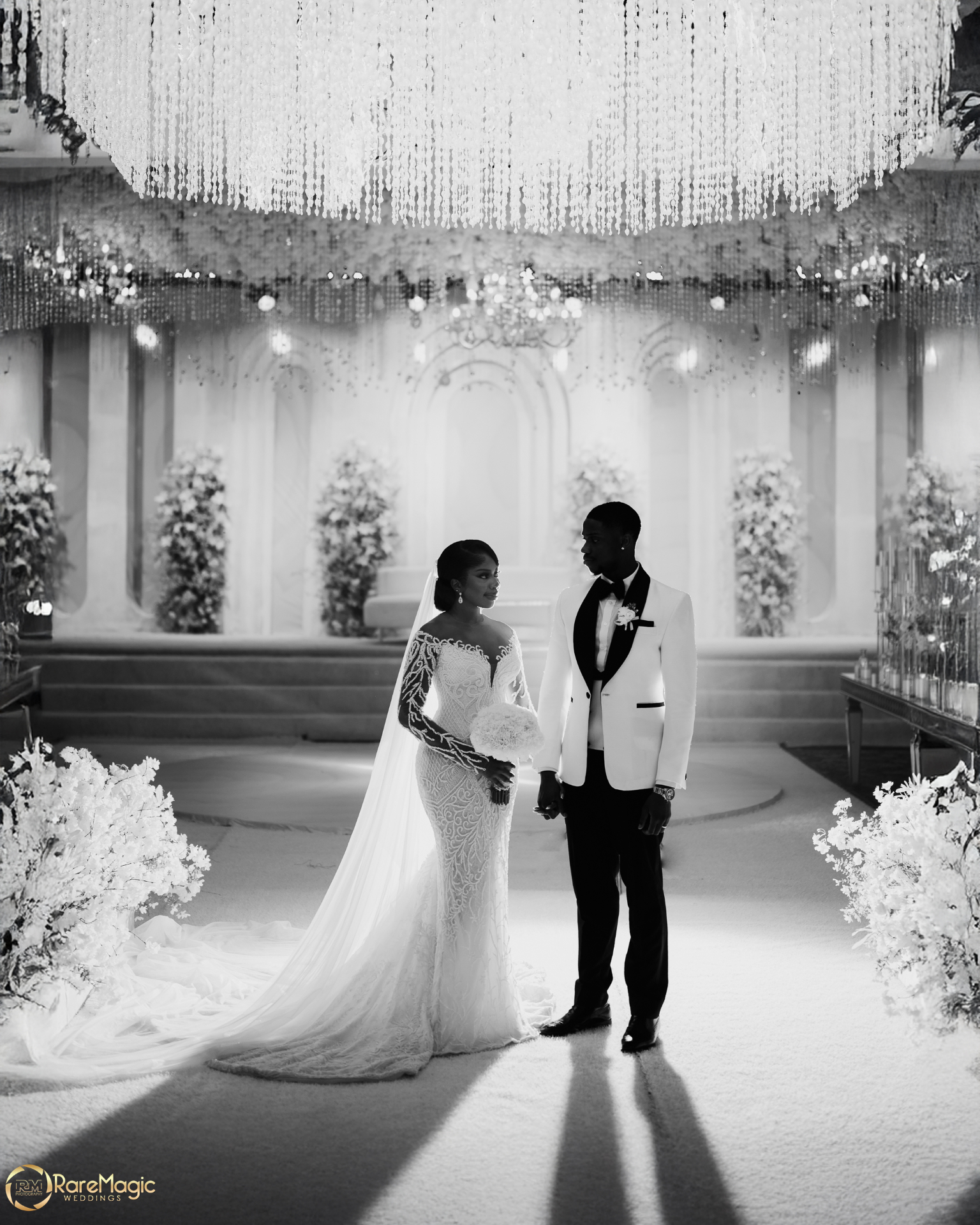 Black and white portrait of the couple standing beneath chandeliers in the reception hall