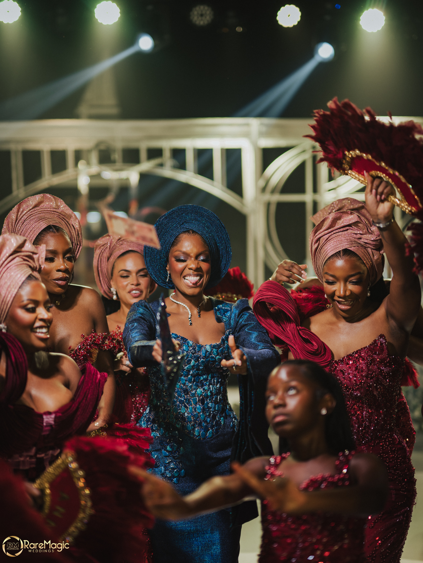 Bride dancing with bridesmaids in coral gele and burgundy gowns under stage lights